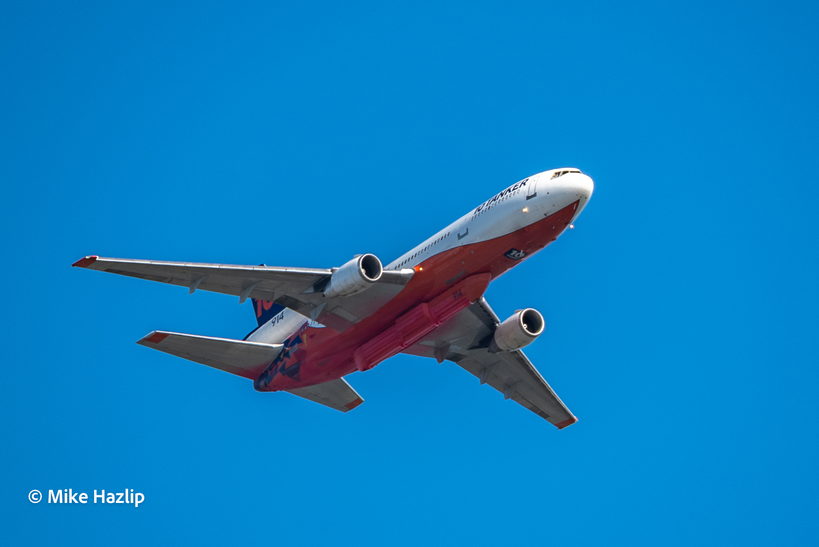 Low-angle photo of a red-and-white DC-10 air tanker against a clear blue sky, "10 TANKER" and tanker number 914 visible on the fuselage.