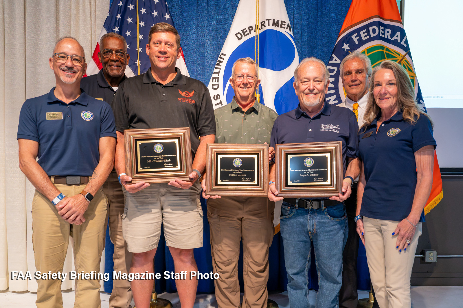 Seven people stand in front of U.S., Department of Transportation and FAA flags. In the center, Mike Kloch, Mike Jesch and Roger Whittier hold framed General Aviation Awards plaques.